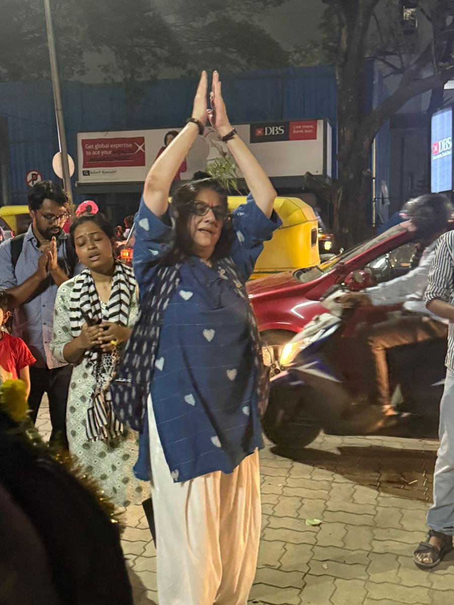 A walker on a Bangalore street at night, hands joined high overhead in kirtan posture, the murti behind